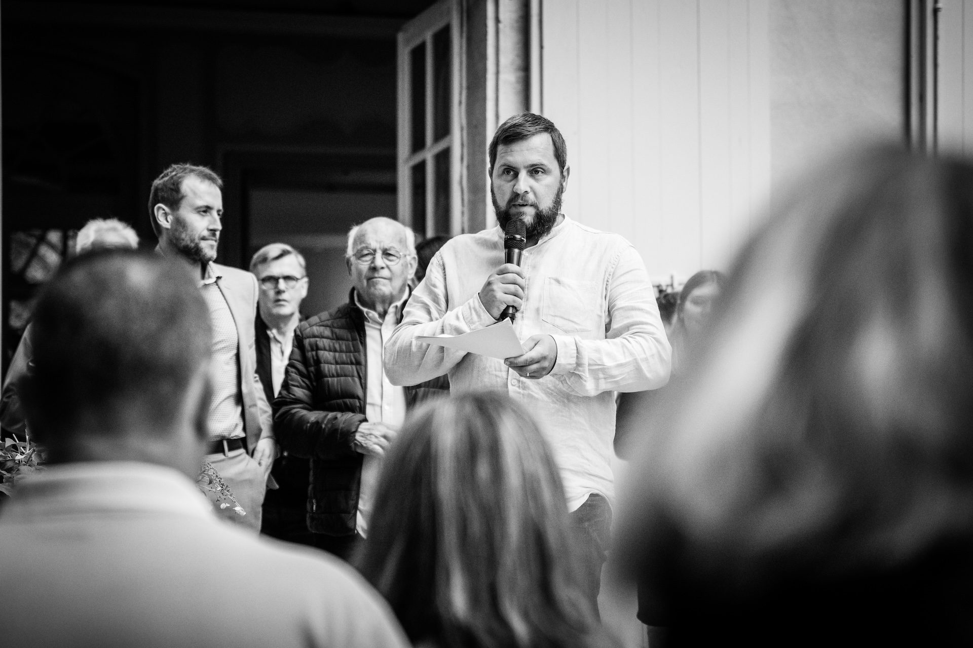 Un invité prenant la parole au micro pendant la garden party, ses notes à la main, entouré d’amis sur le seuil du château ; en noir et blanc