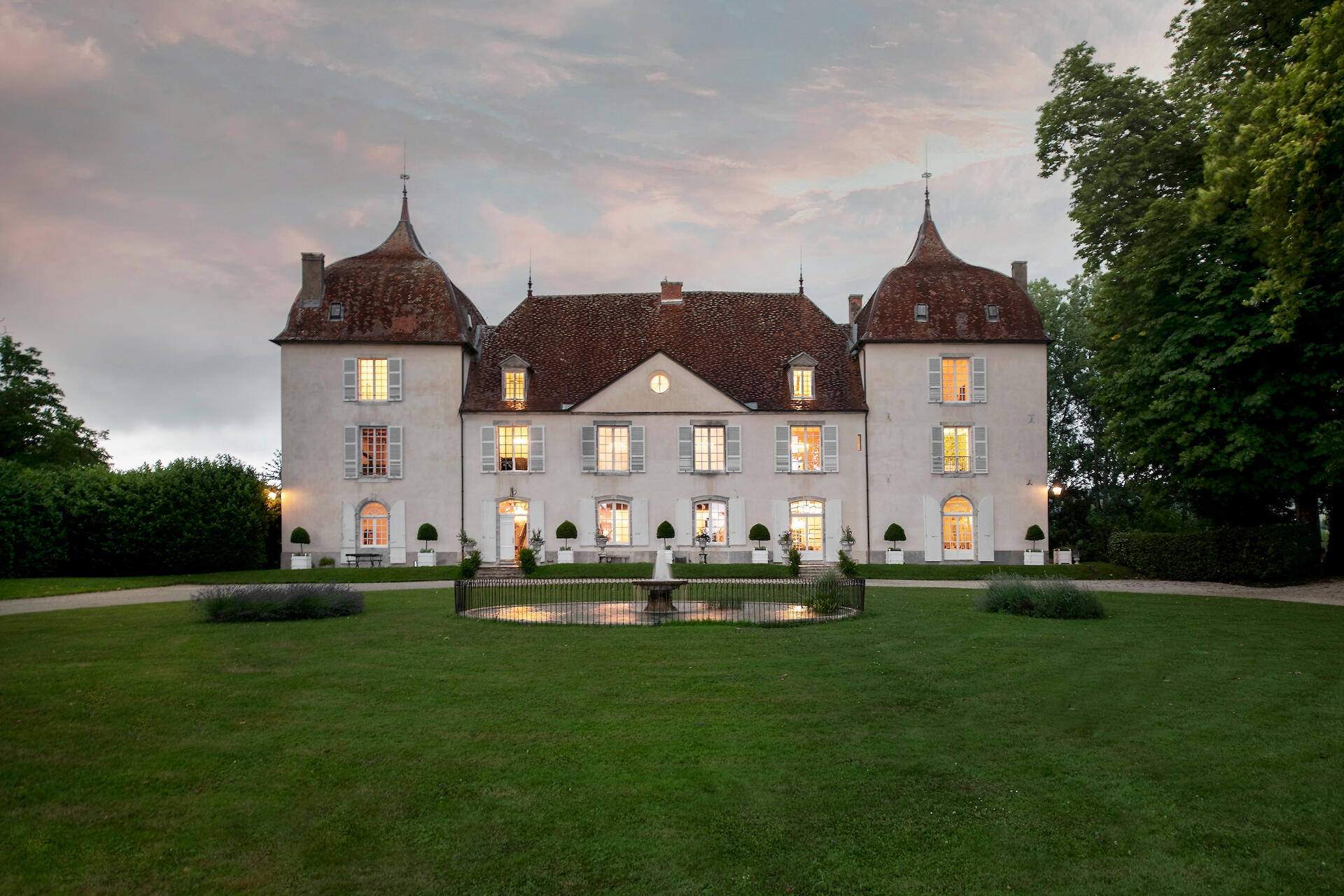 La façade du Château de Roche au crépuscule, ses fenêtres éclairées au-dessus de la pelouse